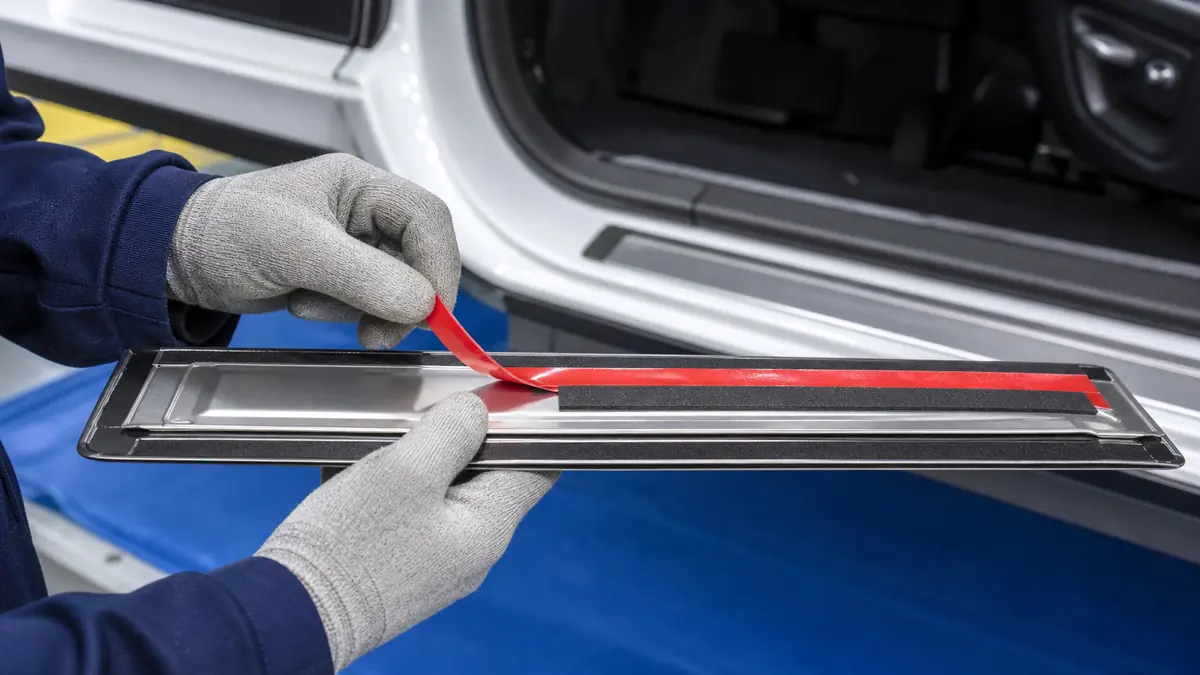 Operator peeling red liner from black double-sided foam tape on an automotive door sill trim plate